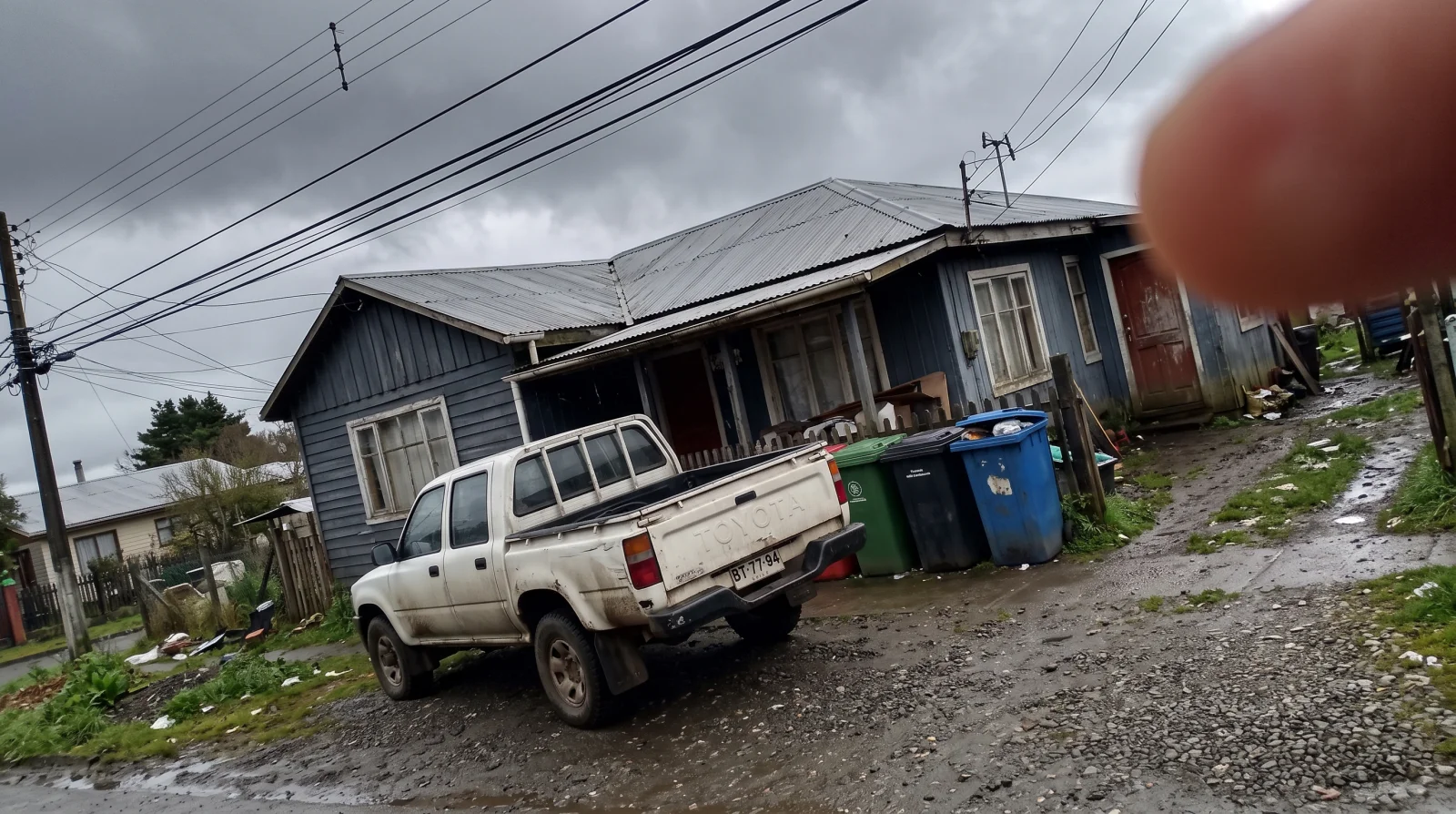 La misma casa fotografiada en un dia gris, con una camioneta tapando la fachada, un dedo sobre el lente, la imagen inclinada y tachos de basura a la vista